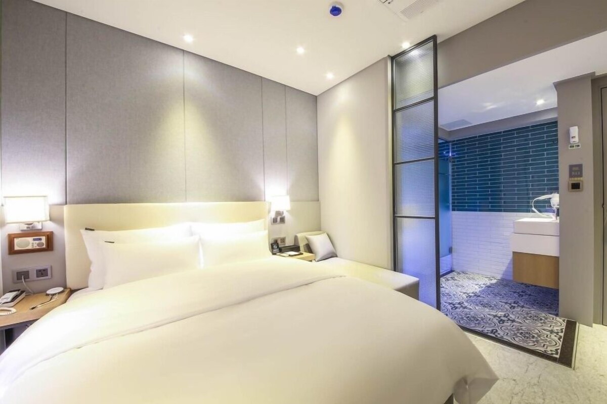 A modern hotel room with a large bed, bedside tables, a nightstand, a bathroom with a glass partition, and a small seating area.