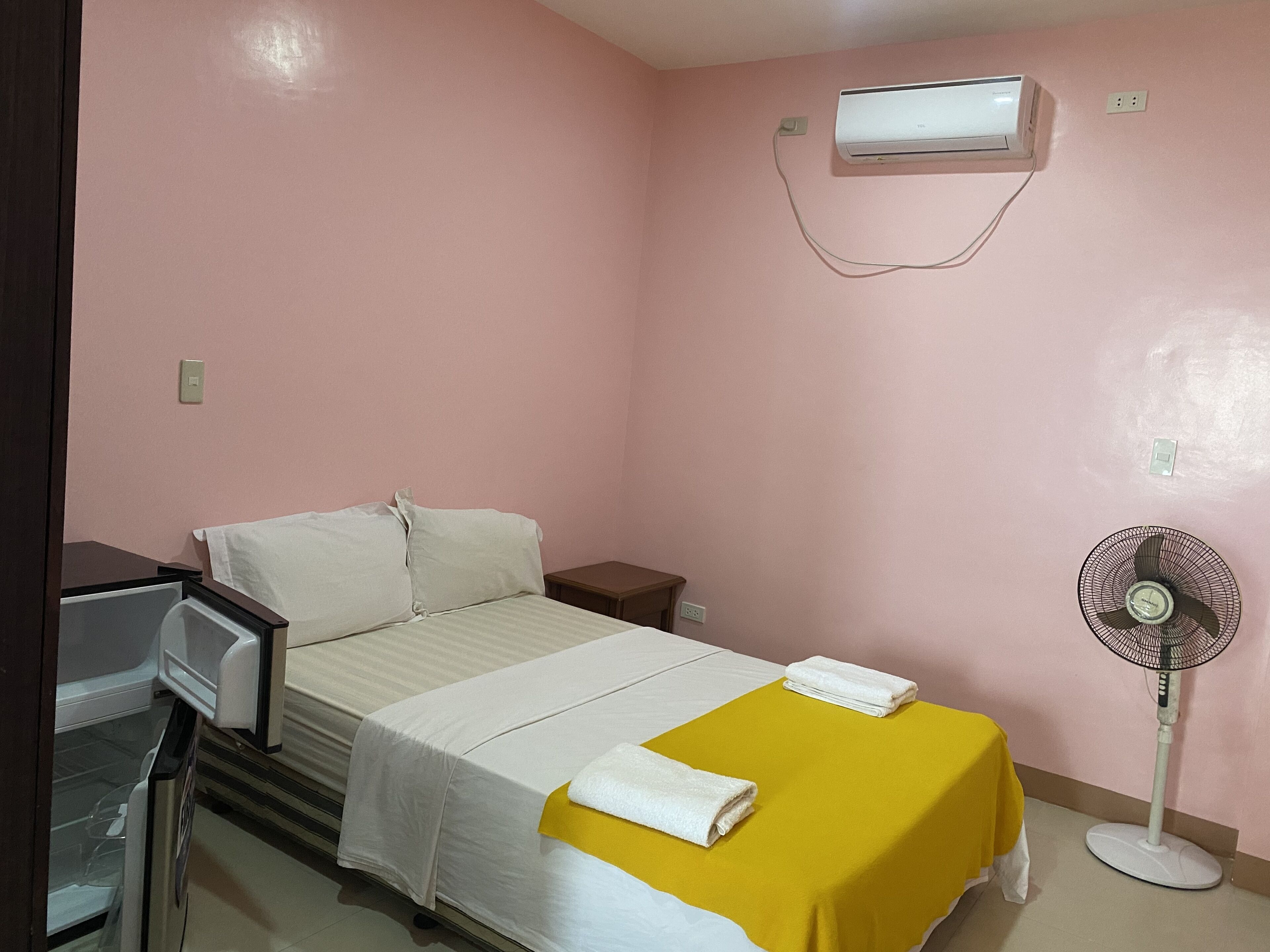 A small, pink-walled room with a bed, a bedside table, a fan, and an air conditioning unit.