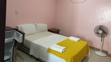 A small, pink-walled room with a bed, a bedside table, a fan, and an air conditioning unit.