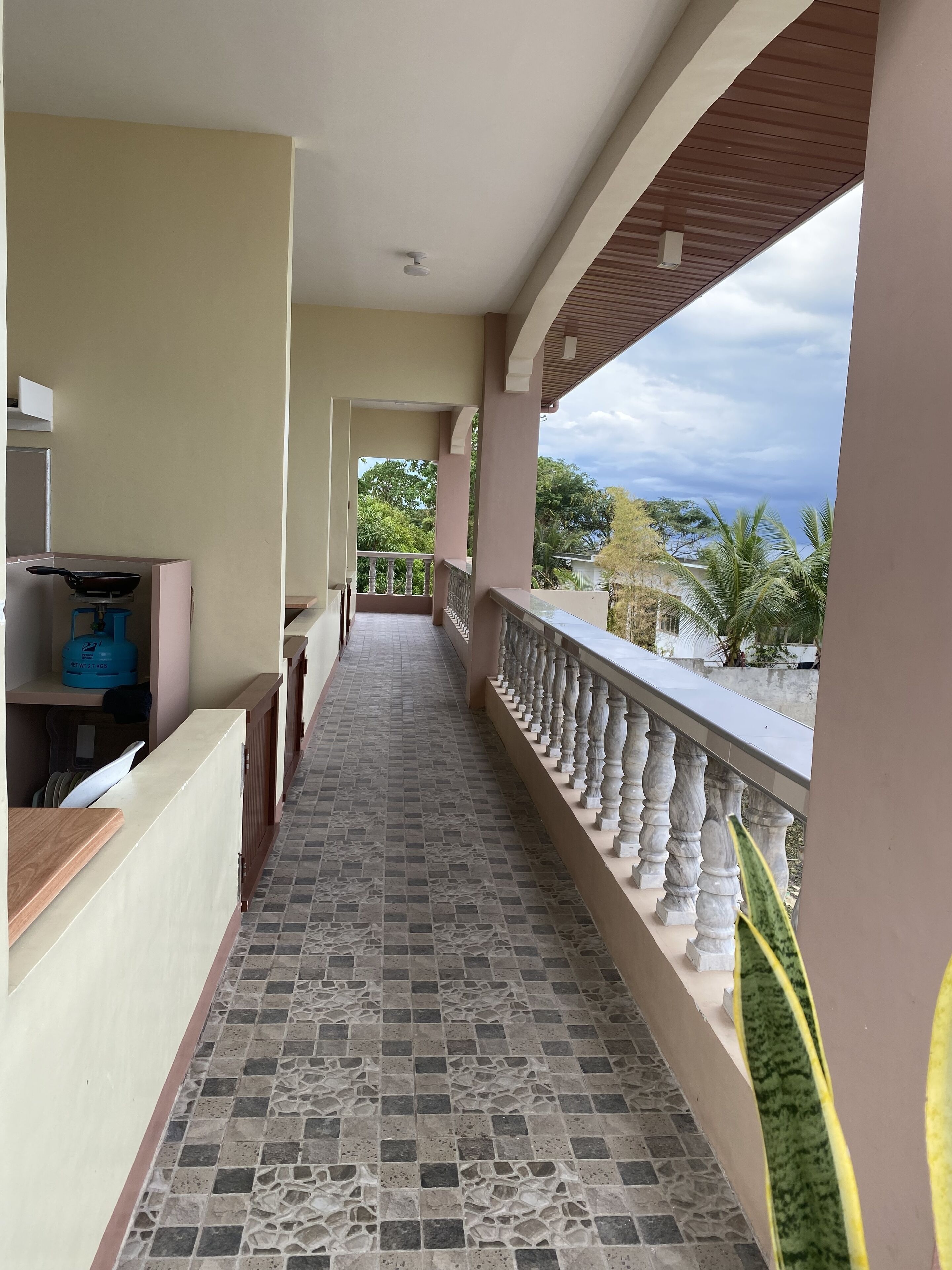 A corridor with a tiled floor, a kitchen area with a stove and blue gas cylinder, and a balcony with a railing and greenery in the background.