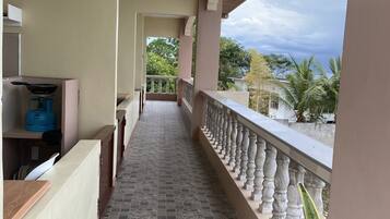 A corridor with a tiled floor, a kitchen area with a stove and blue gas cylinder, and a balcony with a railing and greenery in the background.