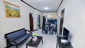 Una sala de estar con sofá, mesa de centro y mueble para la televisión. Hay un cuadro en la pared y una cocina al fondo.