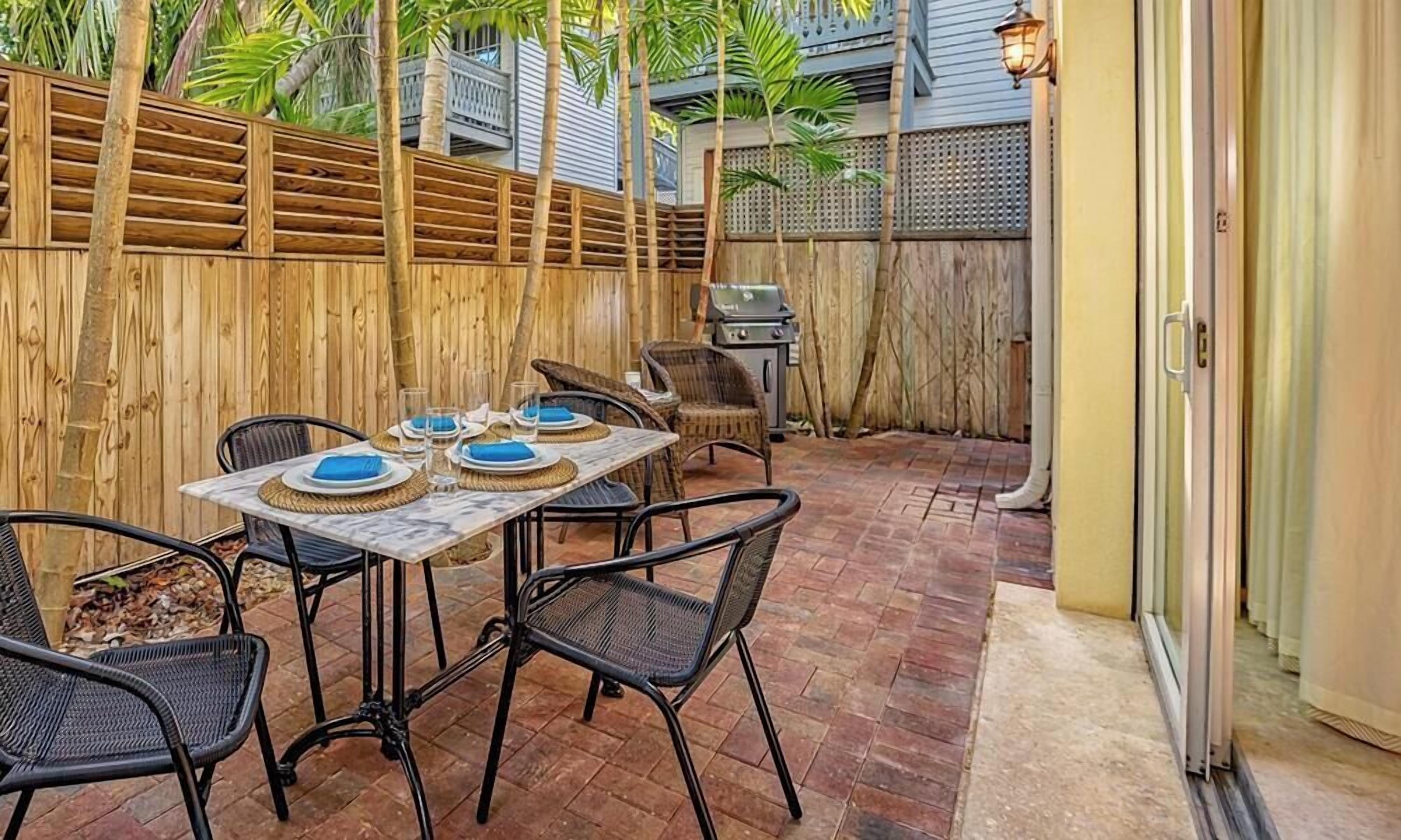 An outdoor patio with a wooden fence, a dining table set with plates and glasses, and wicker chairs.