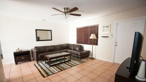A living room with a brown leather sofa, a coffee table, a TV on a stand, a ceiling fan, a window with blinds, and a lamp.