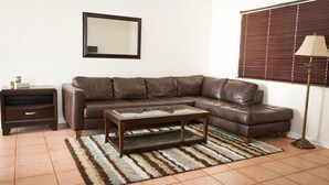 A living room with a brown leather sectional sofa, a glass-top coffee table, a dark wooden side table, a striped rug, a floor lamp, and a window with blinds.