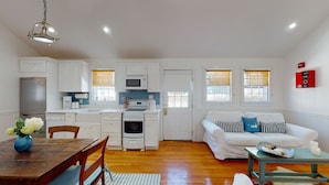 A compact living space with a kitchenette, a dining area with a wooden table and chairs, a white sofa with striped pillows, and a small side table with candles and snacks.