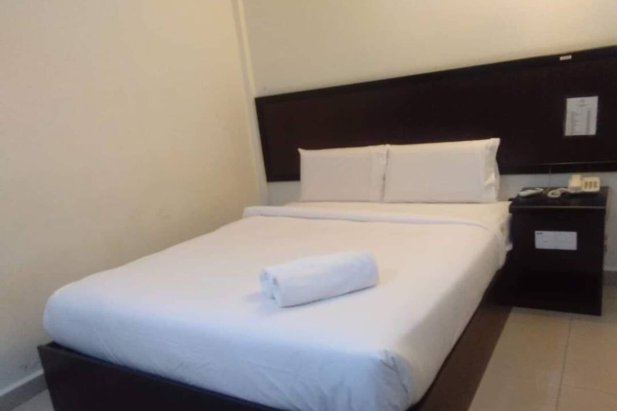 Una cama bien tendida con sábanas blancas y una toalla doblada encima, un cabecero de madera oscura y una mesita de noche con una lámpara y un teléfono.