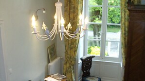 A room with a wooden floor, a patterned rug, a chair, a small table, a window with curtains, and a chandelier. - Chambre d'hôtes - Domaine De Berthegille (Sablonceaux)