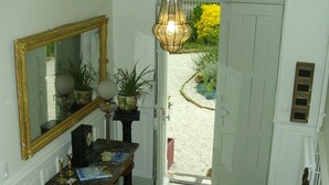 A hallway with a checkered floor, a mirror, a table with a lamp, a chair, and a door leading outside. - Chambre d'hôtes - Domaine De Berthegille (Sablonceaux)
