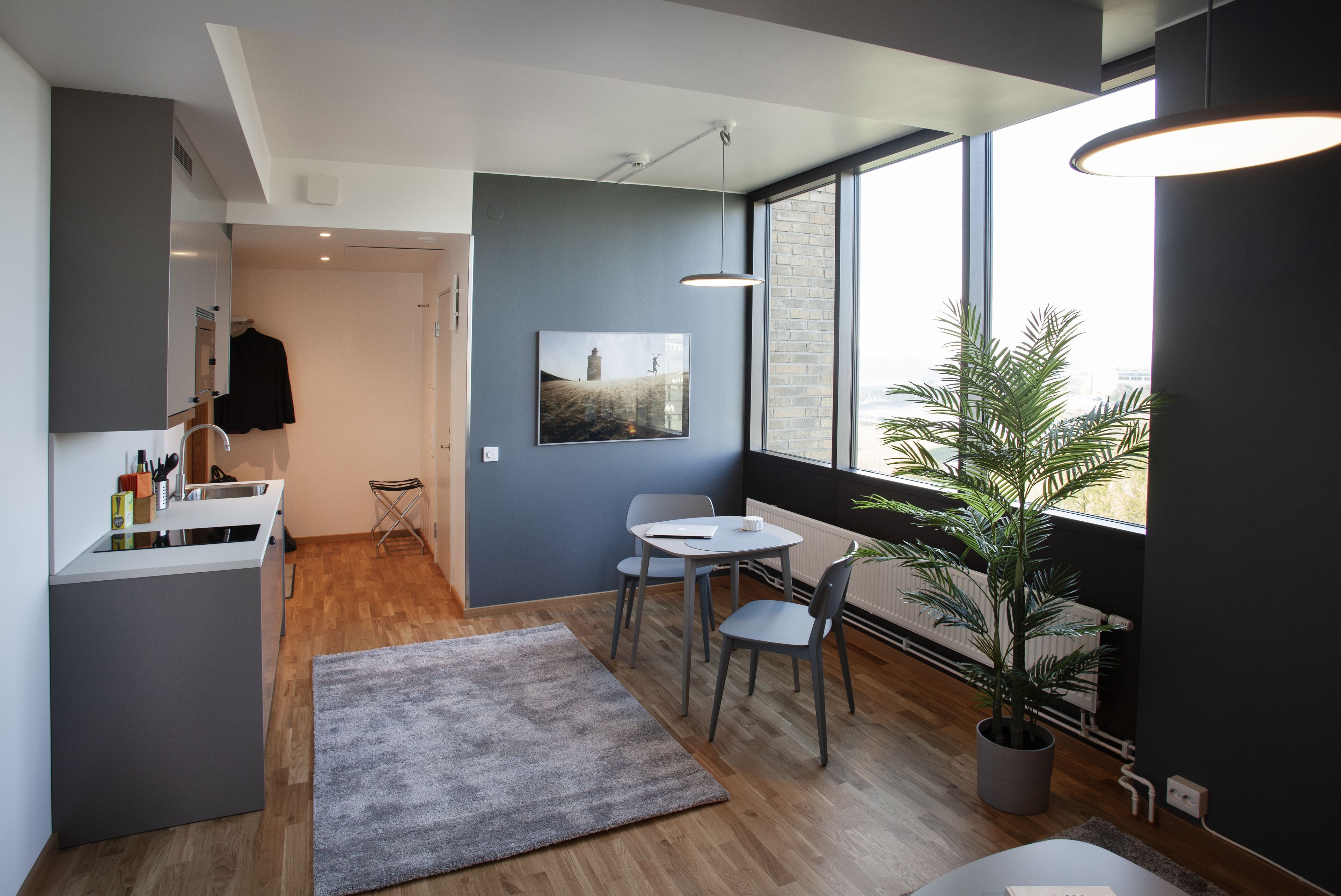 A modern kitchen with a grey countertop, a sink, and a hanging rack. There is a dining area with a table and chairs, and a large window with a view of the city.