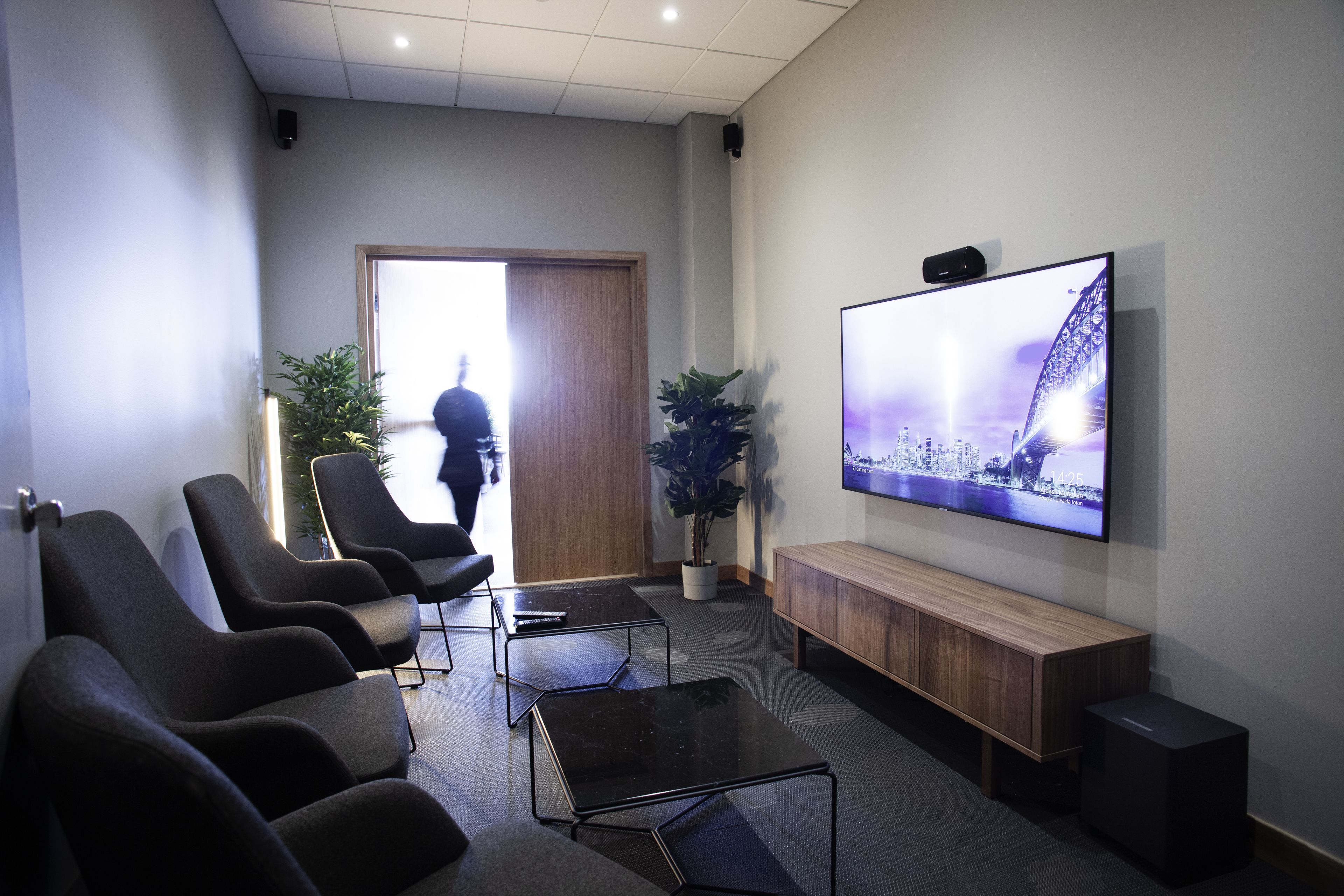 Un área de espera moderna con sillas negras, un banco de madera, un televisor de pantalla plana y una planta en maceta.