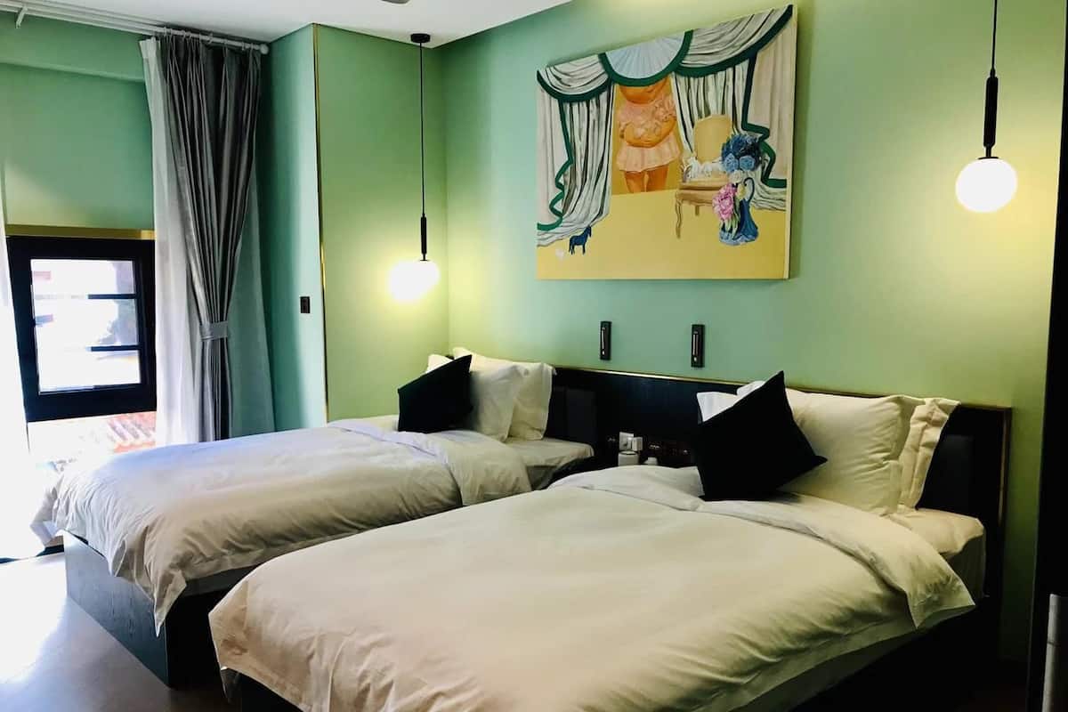 Hotellihuone, jossa on kaksi sänkyä, suuri ikkuna, jossa on verhot, seinällä taulu ja katosta roikkuva valaisin.