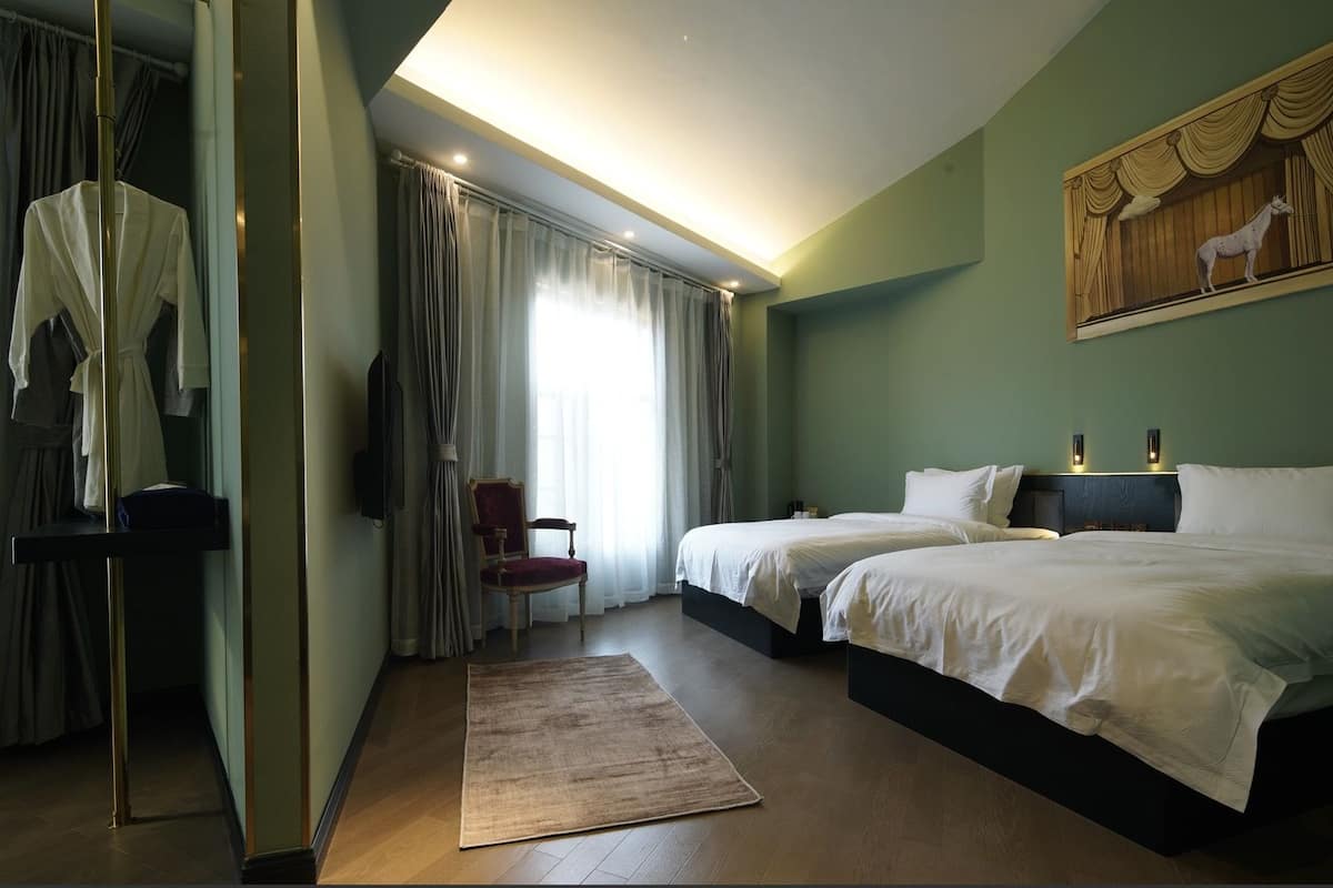 Hotellihuone, jossa on kaksi sänkyä, tuoli, työpöytä ja taulu seinällä.