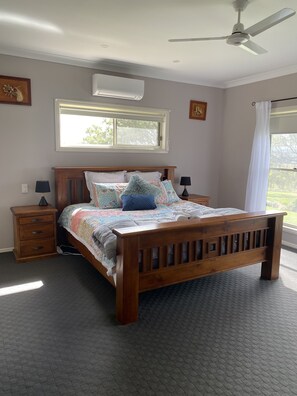 Ein Schlafzimmer mit einem hölzernen Bett, einem Nachttisch, einer Lampe, einem Deckenventilator und einem Fenster mit Blick auf Grünflächen.