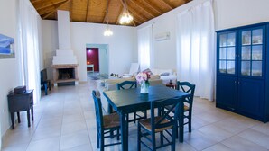 Een ruime woonkamer met houten plafond, een eettafel met stoelen, een blauwe kast en een open haard.