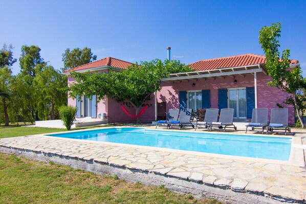 A pink villa with a swimming pool, outdoor seating, and a red roof.