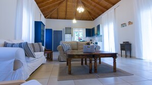 Een ruime woonkamer met houten plafond, een bank, een salontafel en een keuken op de achtergrond.