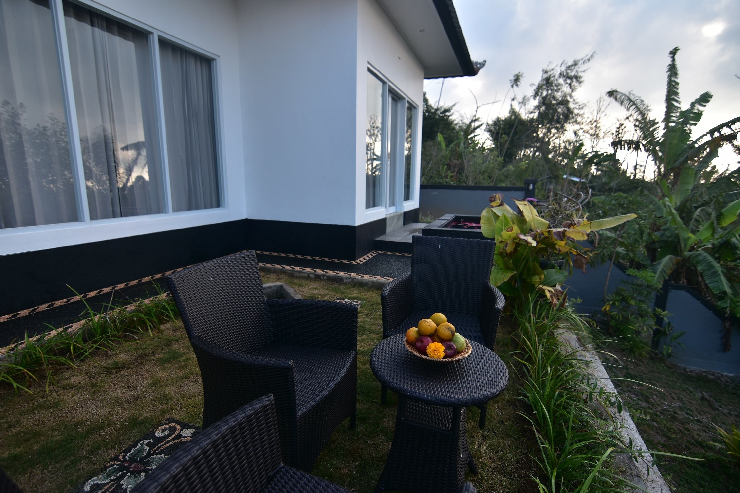 Een modern buitenzitgedeelte met rieten stoelen en een kleine tafel, een fruitschaal en een huis met grote ramen en een tuin.