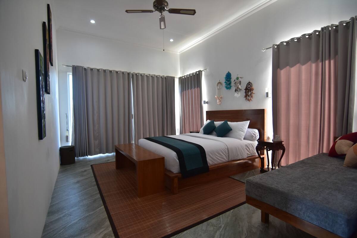 A bedroom with a large bed, a smaller bed, a wooden bench, a ceiling fan, and a view of the outside.