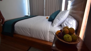 A neatly made bed with white linens and a teal pillow, a wooden nightstand with a fruit basket, and a window with curtains. - Wanagiri Cosmic Nature Villa (Wanagiri)