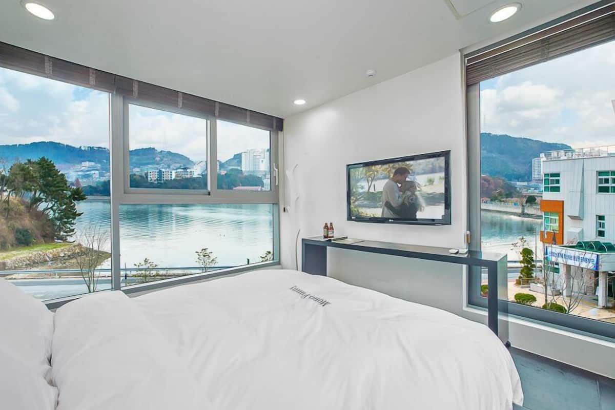 A hotel room with a large window offering a scenic view of a lake and buildings, a bed with white linens, and a flat-screen TV mounted on the wall.