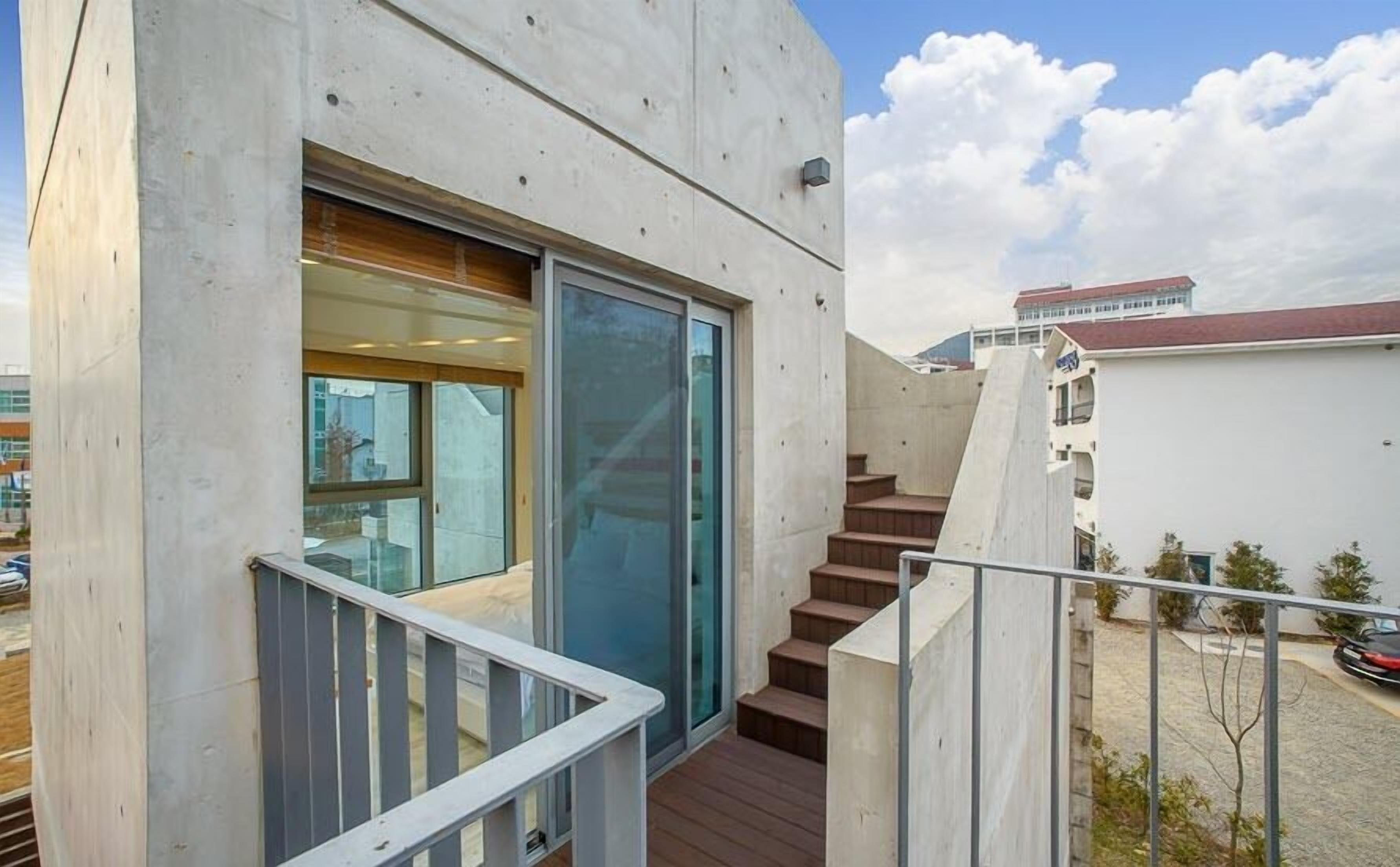 A modern building with a concrete facade, a glass door, and a wooden staircase leading to a deck.