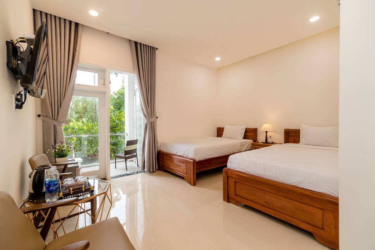 A hotel room with two beds, a desk with a chair, a TV, and a balcony with curtains.