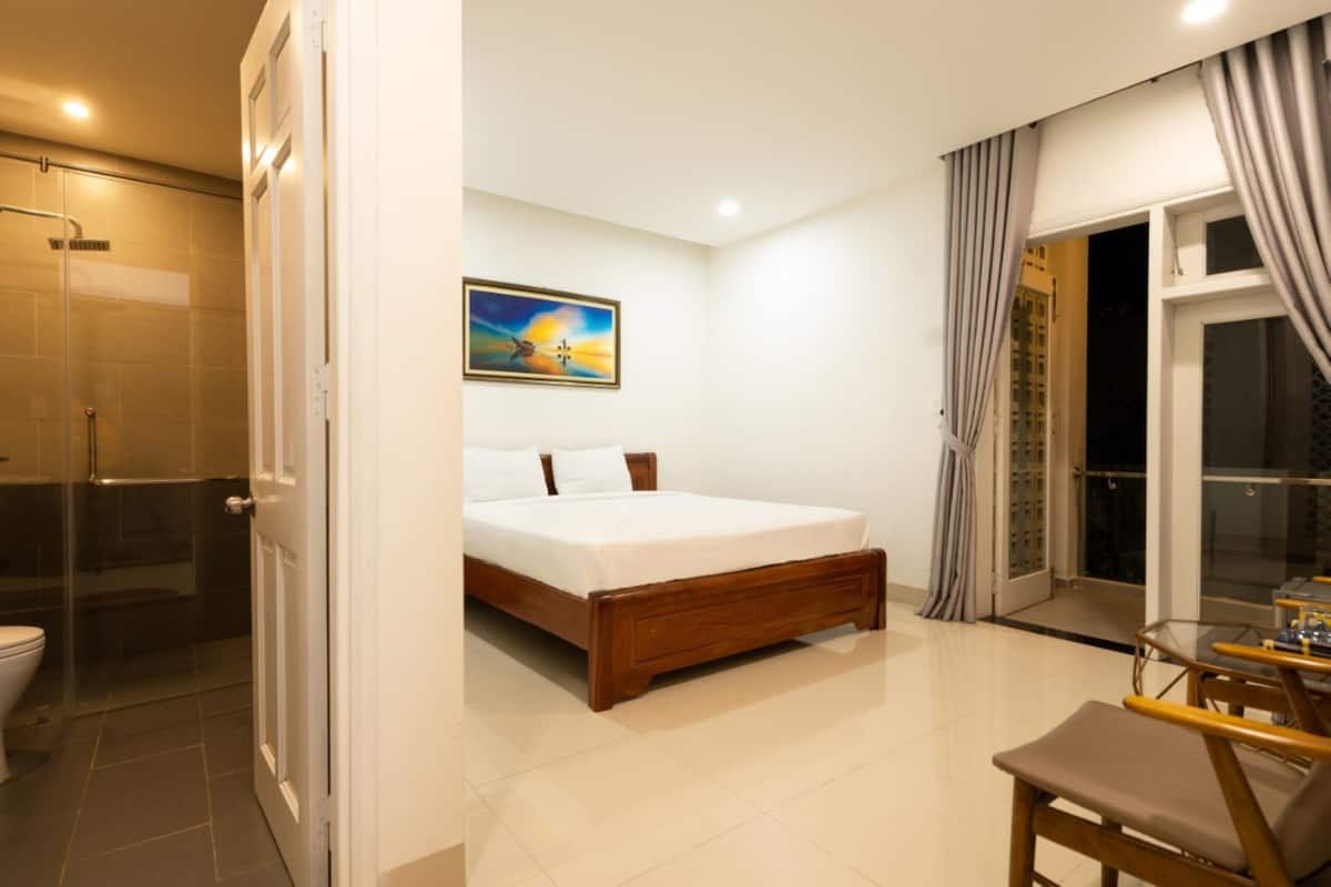 A bedroom with a bed, a painting, a bathroom with a shower, and a balcony with curtains.
