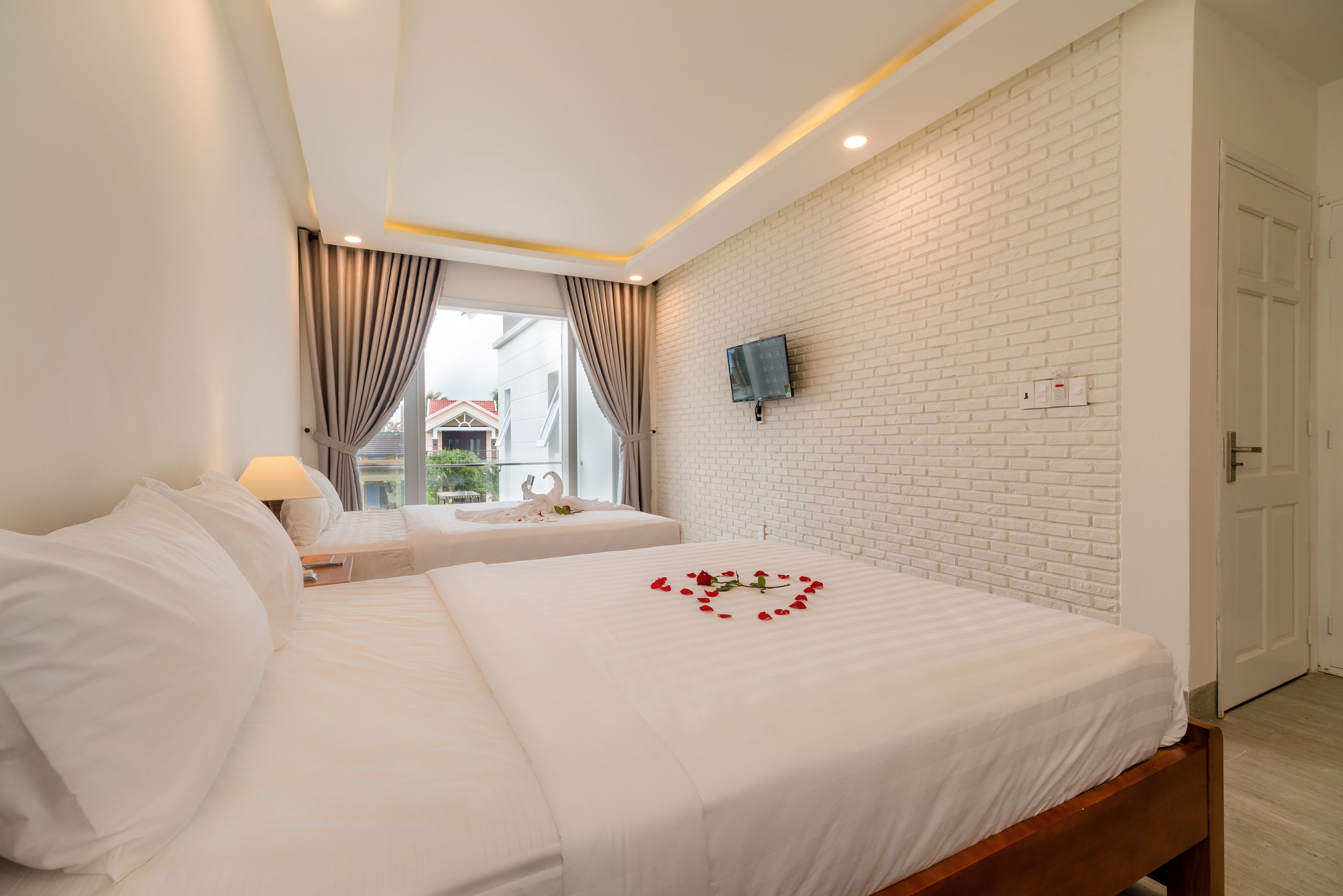 A hotel room with a white brick wall, a flat-screen TV mounted above the bed, and a window with curtains.