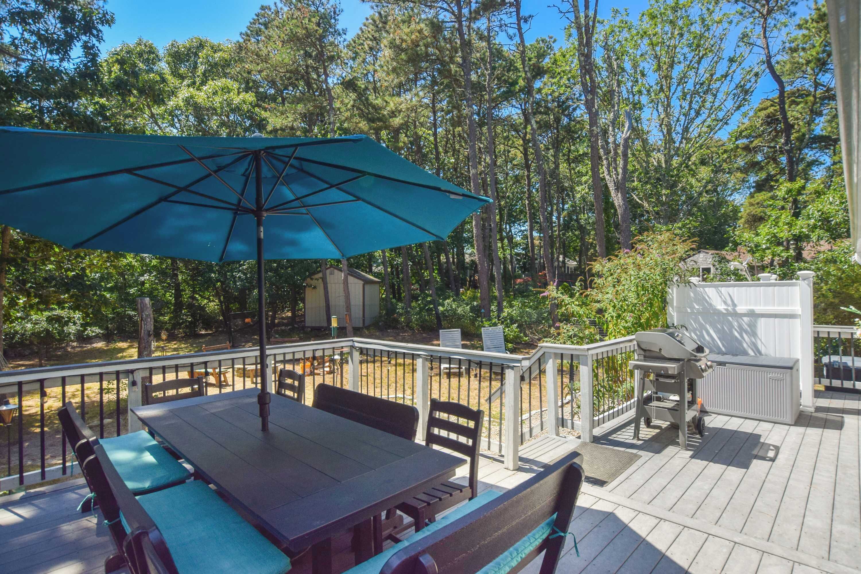 A wooden deck with a table and chairs, a blue umbrella, and a barbecue grill.