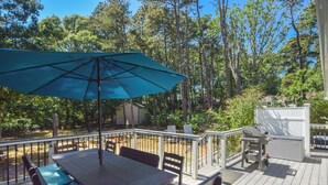Un terrazzo in legno con tavolo e sedie, un ombrellone blu e un barbecue.