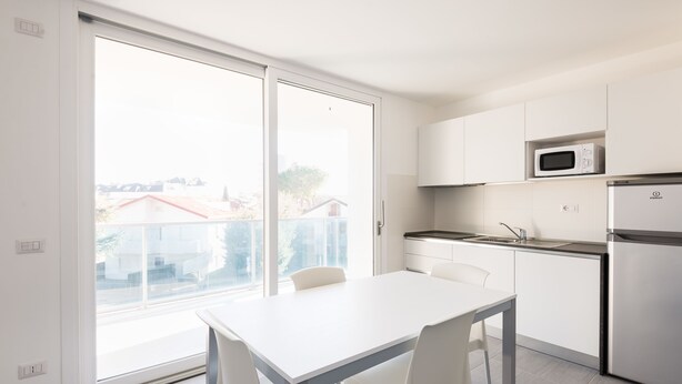 Een moderne keuken met witte kasten, een magnetron en een koelkast. Er staat een eettafel met stoelen en een schuifpui naar een balkon.