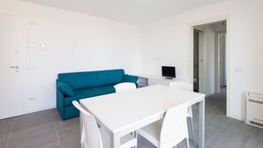 Een moderne, minimalistische kamer met een witte tafel en stoelen, een blauwgroene bank en een flatscreen-televisie.
