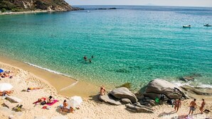 A beach with clear turquoise water, people sunbathing, and rocky formations. - Faro di Punta Fenaio (Isola del Giglio)