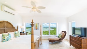 A bedroom with a large bed, a ceiling fan, a TV, a wicker chair, and a view of the ocean.