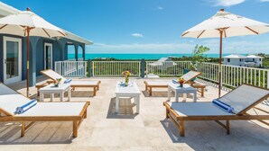 A sunlit outdoor seating area with lounge chairs and tables, overlooking a turquoise sea.