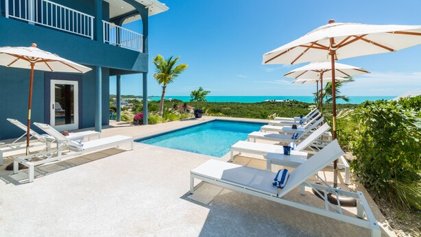 A pool area with lounge chairs and umbrellas, a clear view of the ocean, and a two-story building with balconies.