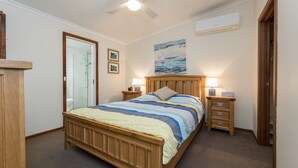 A bedroom with a wooden bed, two bedside tables, a nightstand, a ceiling fan, a shower, and a painting on the wall. - Inlet Dreaming - Inlet Side Inverloch (INVERLOCH)