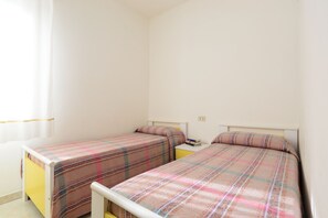 A bedroom with two single beds, each with a plaid bedspread, a bedside table with a book, and a plain wall with a light switch.