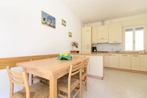 Een eetgedeelte met een houten tafel en stoelen, een keuken met lichte kastjes, en een klein eilandje met een bloevas.