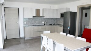 A modern kitchen with a wooden table and white chairs, a refrigerator, and a microwave. - Appartamenti Nashira (Lignano Sabbiadoro)