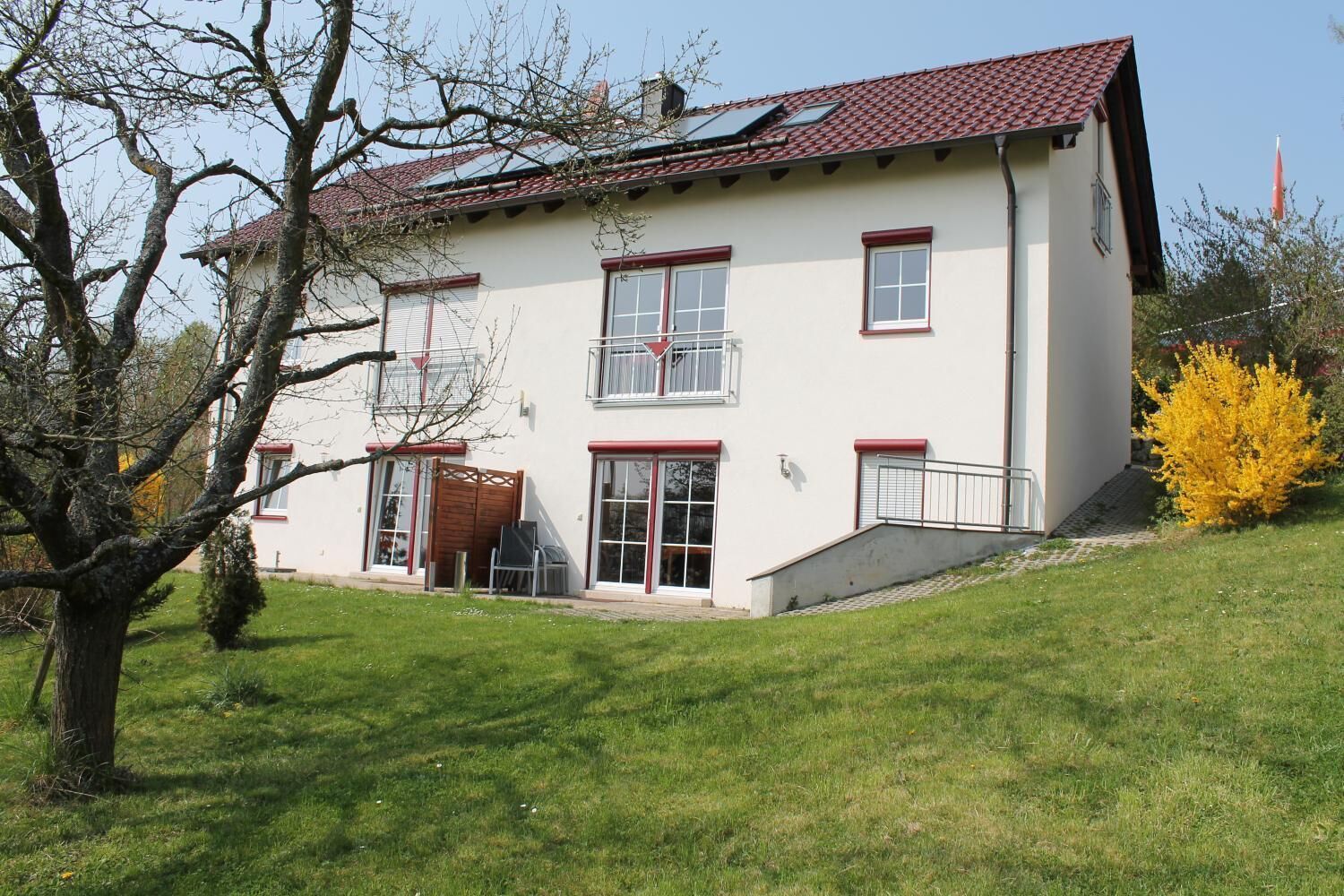 A two-story house with a red-tiled roof, white walls, and red-framed windows. There is a small front porch and a staircase leading to the entrance.