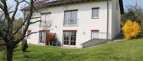 A two-story house with a red-tiled roof, white walls, and red-framed windows. There is a small front porch and a staircase leading to the entrance.