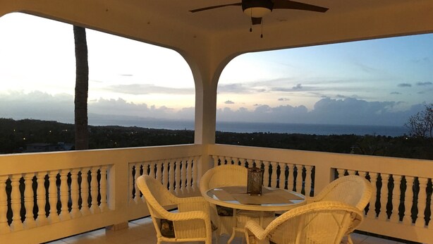 A covered balcony with a round table and four wicker chairs, overlooking a scenic landscape with mountains and a body of water. - Casa Anonna - Stunning oceans views and pool! (Vieques)