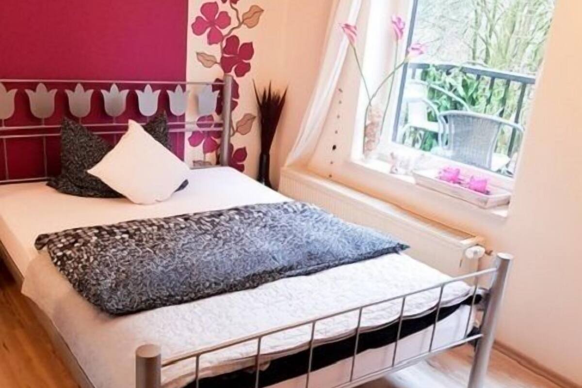 A bedroom with a metal bed frame, a window with a view of greenery, and a pink wall with floral wallpaper.