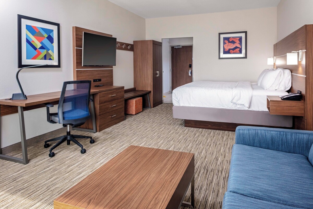 A hotel room with a bed, a desk with a chair, a TV, and artwork on the walls.