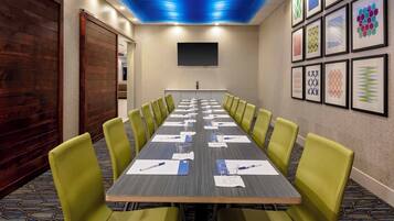A conference room with a long table, green chairs, a projector screen, and framed artwork on the walls.