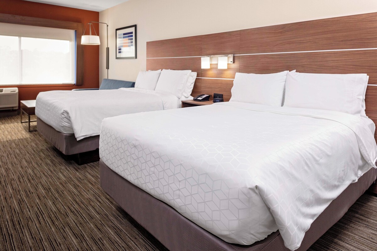 A hotel room with two beds, a desk, a chair, a window, and a wall-mounted lamp.