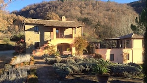A two-story stone house with a balcony, surrounded by a landscaped garden with various plants and shrubs. - Casa Rosa (Assisi)