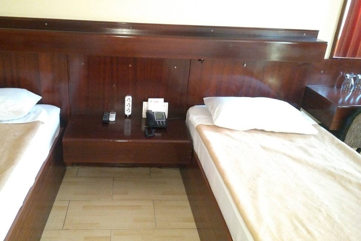 Camera d'albergo con due letti singoli, una testiera in legno, un tavolino con un telefono e una tazza, e un pavimento piastrellato.